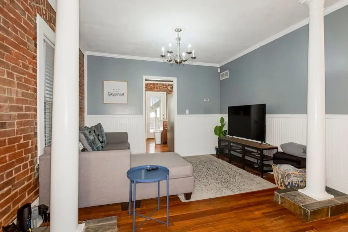 A modern living space showcasing a comfortable sofa, a large television mounted on a brick wall, a chandelier overhead, and tasteful decorative elements including a plant. The room radiates a cozy, welcoming vibe ideal for entertainment or winding down after a busy day.