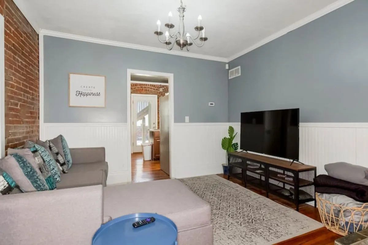 An open-plan living room with a comfortable sofa facing a television, a modern chandelier, and prominent wall artwork. The area is designed for group activities—whether socializing, watching TV, or sharing meals—reflecting a lively, communal atmosphere.