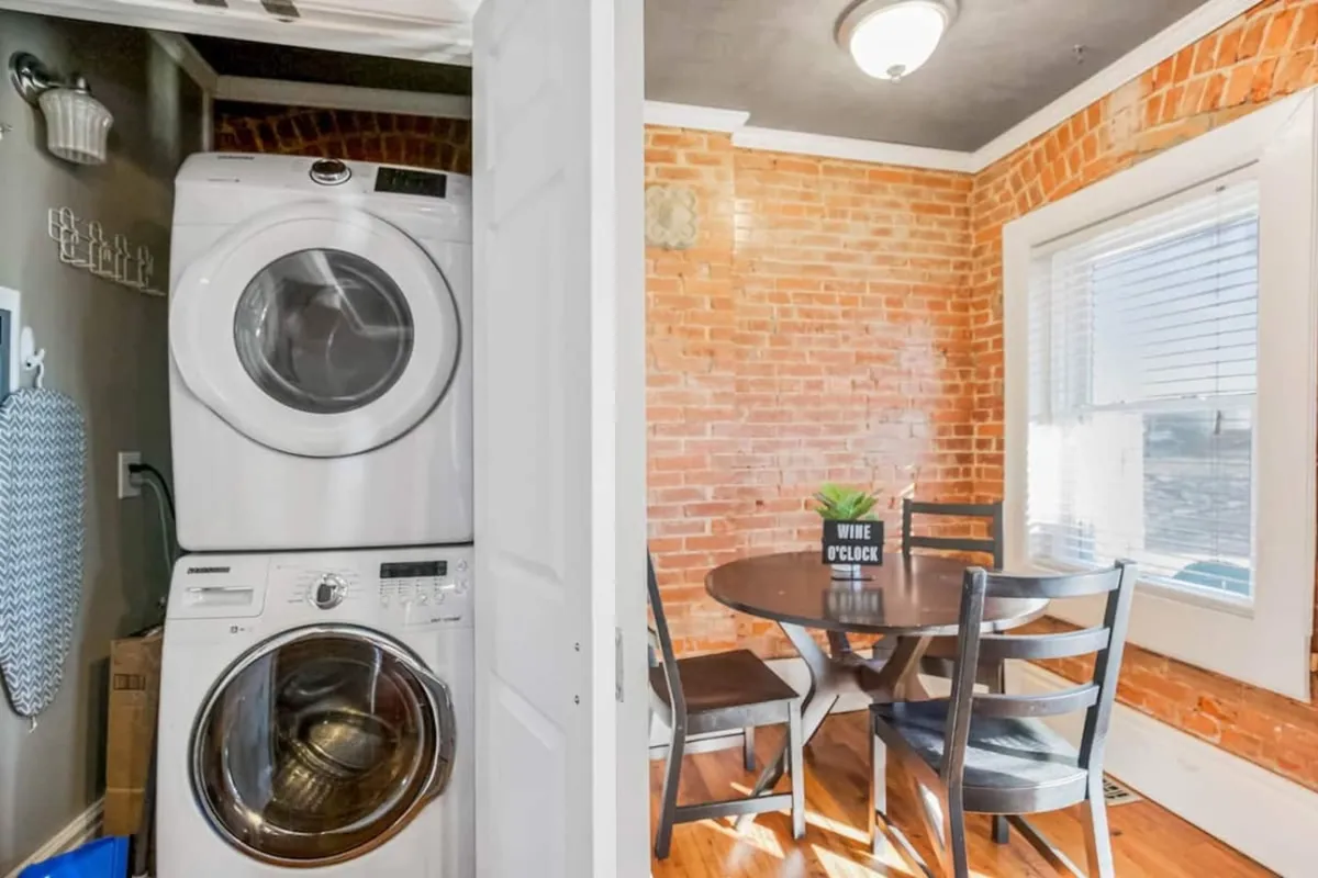 The image features a stacked washer and dryer set against a brick wall, with natural light streaming in from a window. A round dining table is positioned nearby, creating a practical and inviting atmosphere. The setting is both functional and homey, highlighting the convenience of doing laundry within the comfort of the living space.
