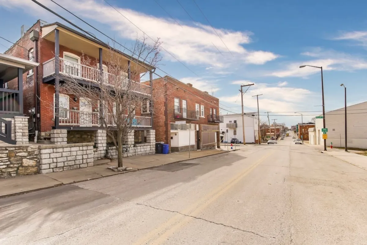 A vibrant streetscape featuring two-story brick buildings with balconies, street trees lining a paved road, and people walking or socializing outdoors. The image exudes a sense of community and the lively, walkable character of the neighborhood.