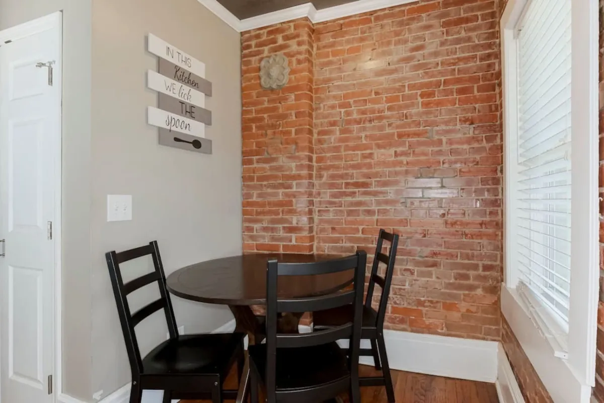 A cozy dining nook is set against an exposed brick wall, featuring a round table and stylish decor, including a kitchen-themed sign. The inviting setting is perfect for intimate meals, morning coffee, or casual gatherings, complementing the kitchen area.