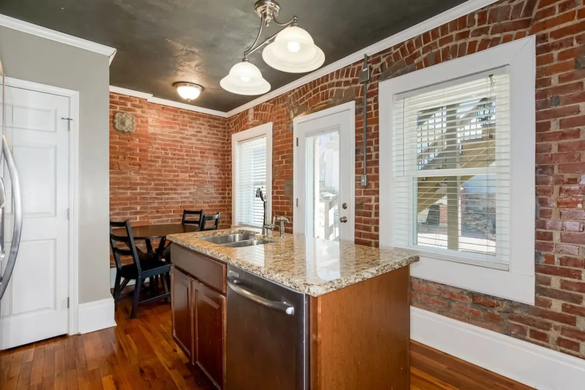 A broader view of the open-plan kitchen and dining area, featuring an expansive granite countertop, exposed brick walls, a kitchen island, large windows, and a wooden dining table. The layout encourages cooking and socializing in a bright, welcoming space.
