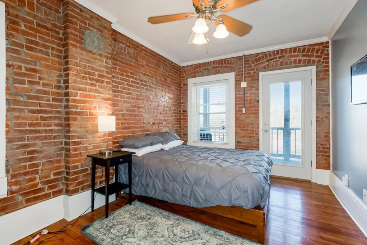A serene bedroom featuring exposed brick walls, a cozy bed with gray bedding, a nightstand with lamp, and a door leading out to a sun-drenched balcony. The fresh linens and thoughtful layout evoke a calm, restful mood ideal for unwinding.