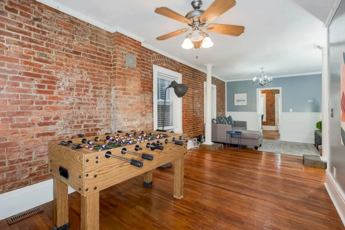 The image showcases a stylish living area with a vibrant foosball table as the centerpiece, set against an exposed brick wall. A comfortable sofa and ceiling fan add to the welcoming atmosphere, creating a space perfect for gathering, relaxing, and sharing laughter.