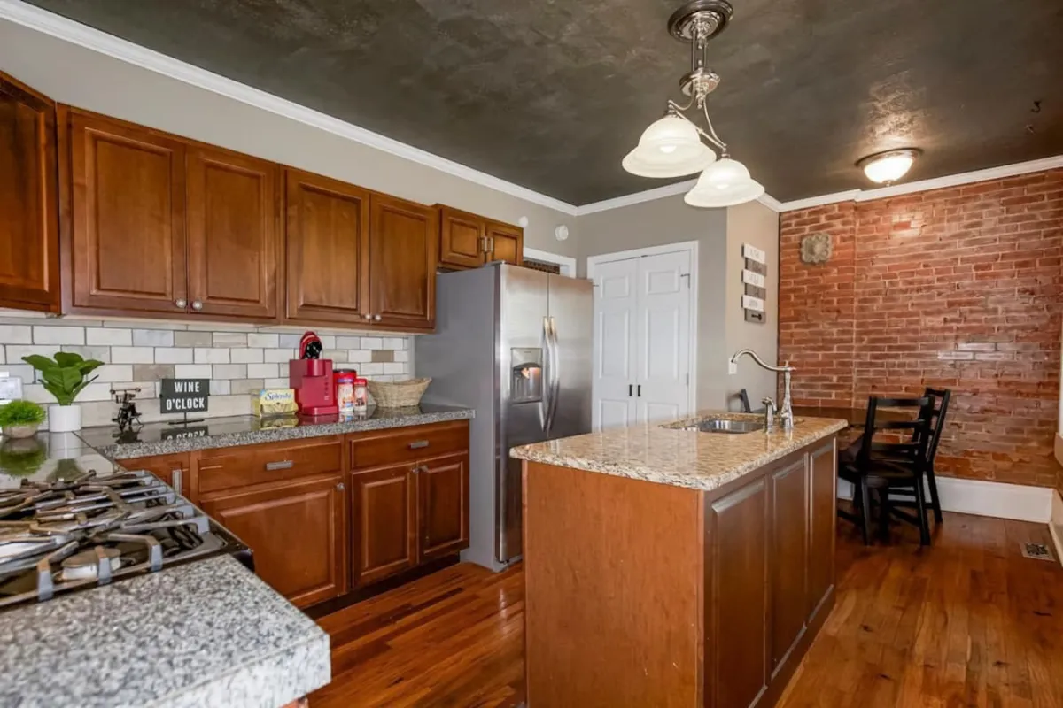 This image showcases a spacious kitchen with granite countertops, stainless steel appliances, a striking exposed brick wall, and wooden cabinetry. The dining area with a chair is visible adjacent to the kitchen, emphasizing an open-concept layout ideal for preparing meals and gathering for dining.